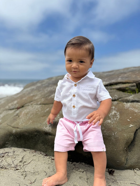 Child in a white shirt and pink shorts standing on a rocky beach with ocean and sky in the background