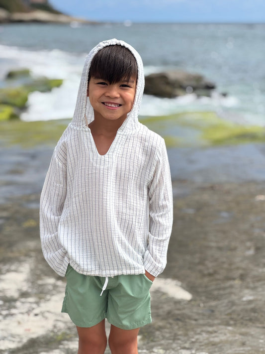 Child wearing a white hooded shirt and green shorts standing on a beach with ocean in the background