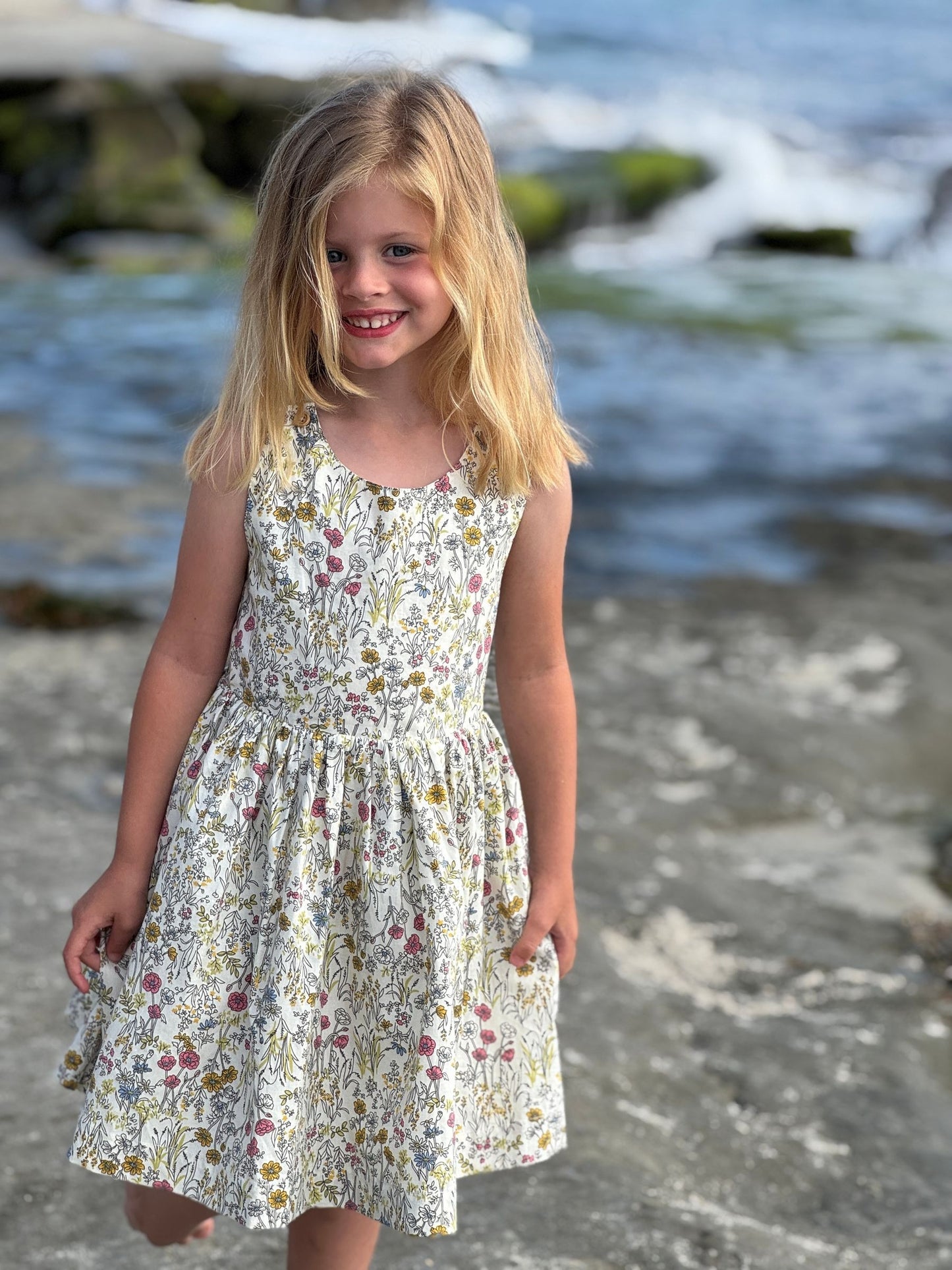 Young girl in a floral dress standing on a rocky beach with ocean waves in the background.