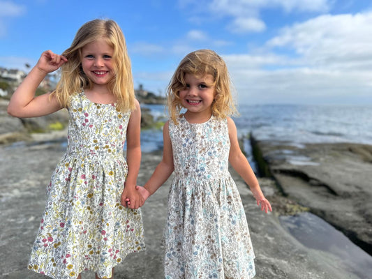 Two young girls in floral dresses standing on a beach with ocean and sky in the background.