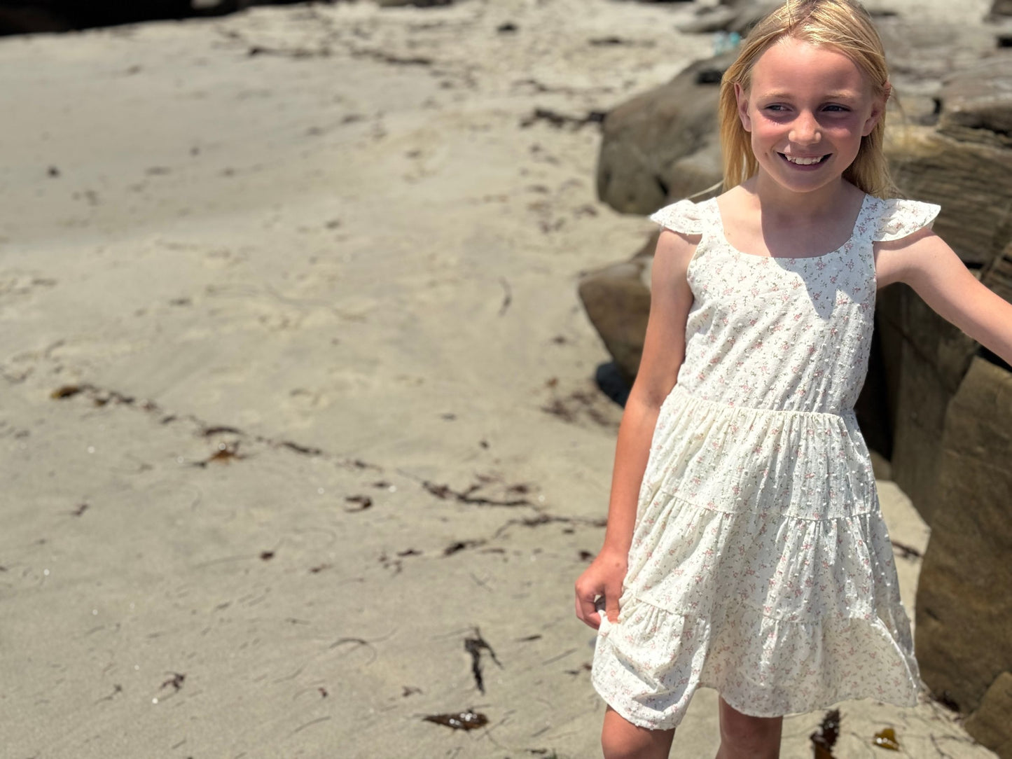 Young girl in a white dress standing on a sandy beach with rocks.
