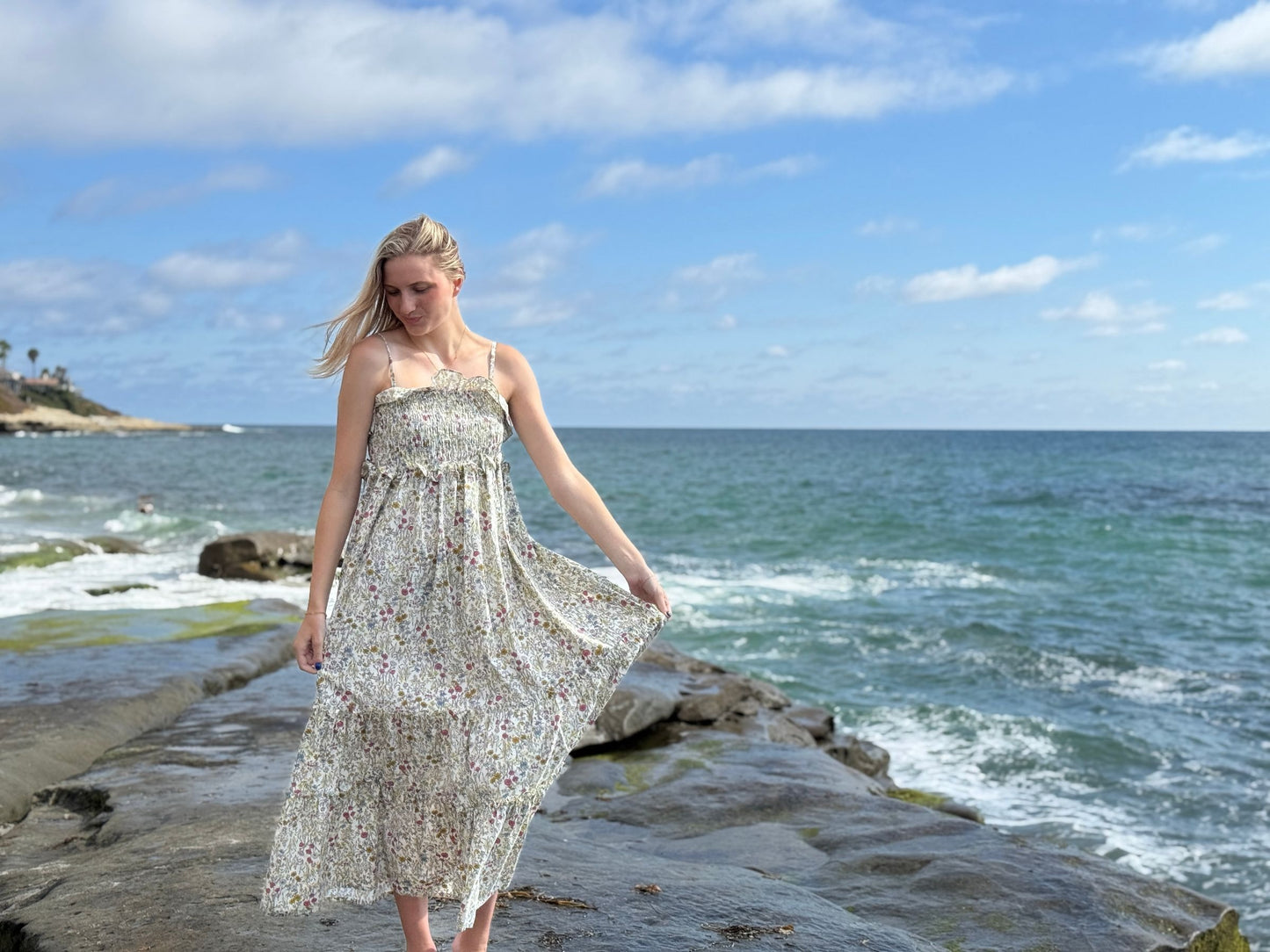 Woman in a floral dress standing on a rocky beach with ocean and sky in the background