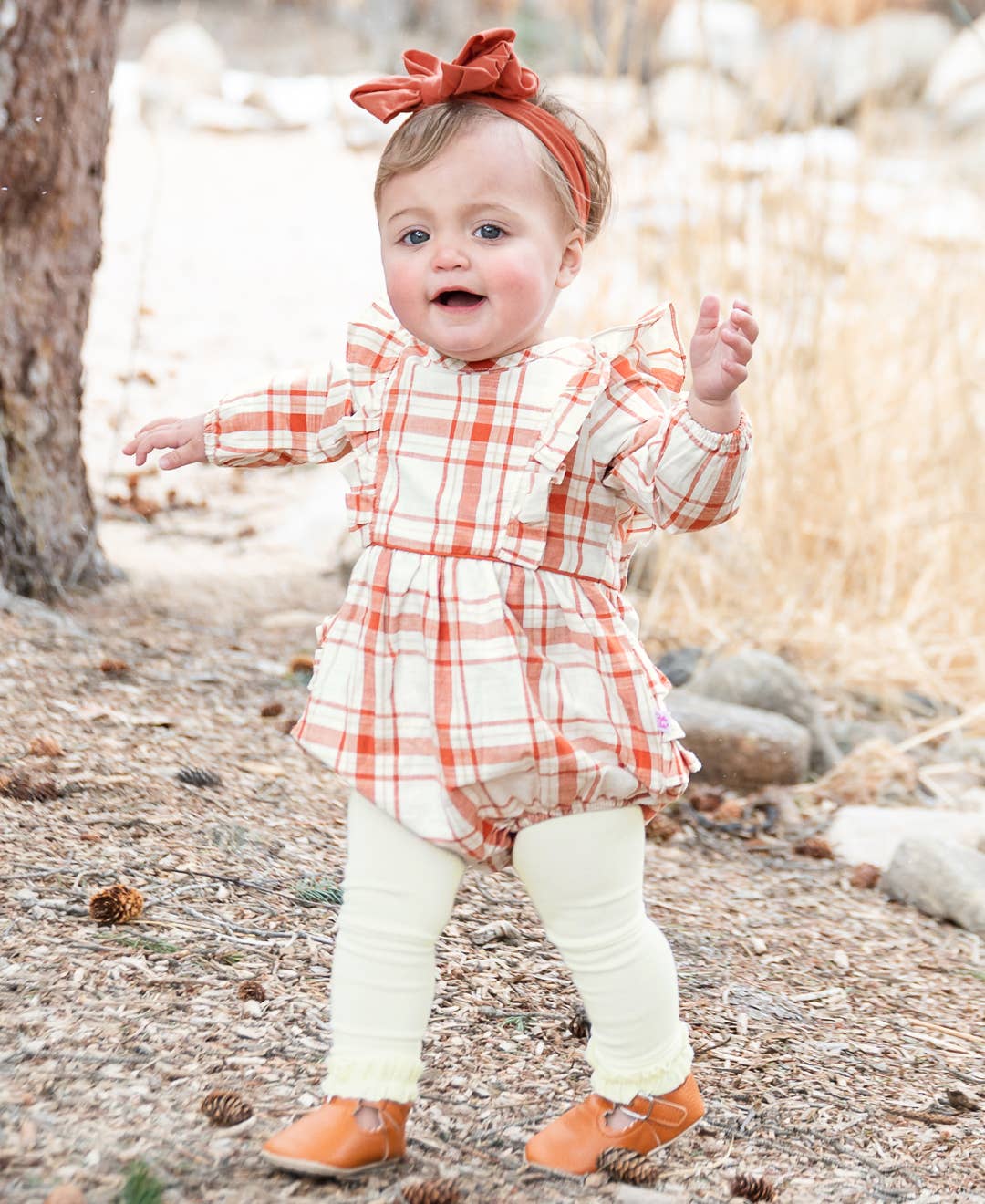 Baby in a red and white checkered dress with a headband, standing outdoors on a rocky path.