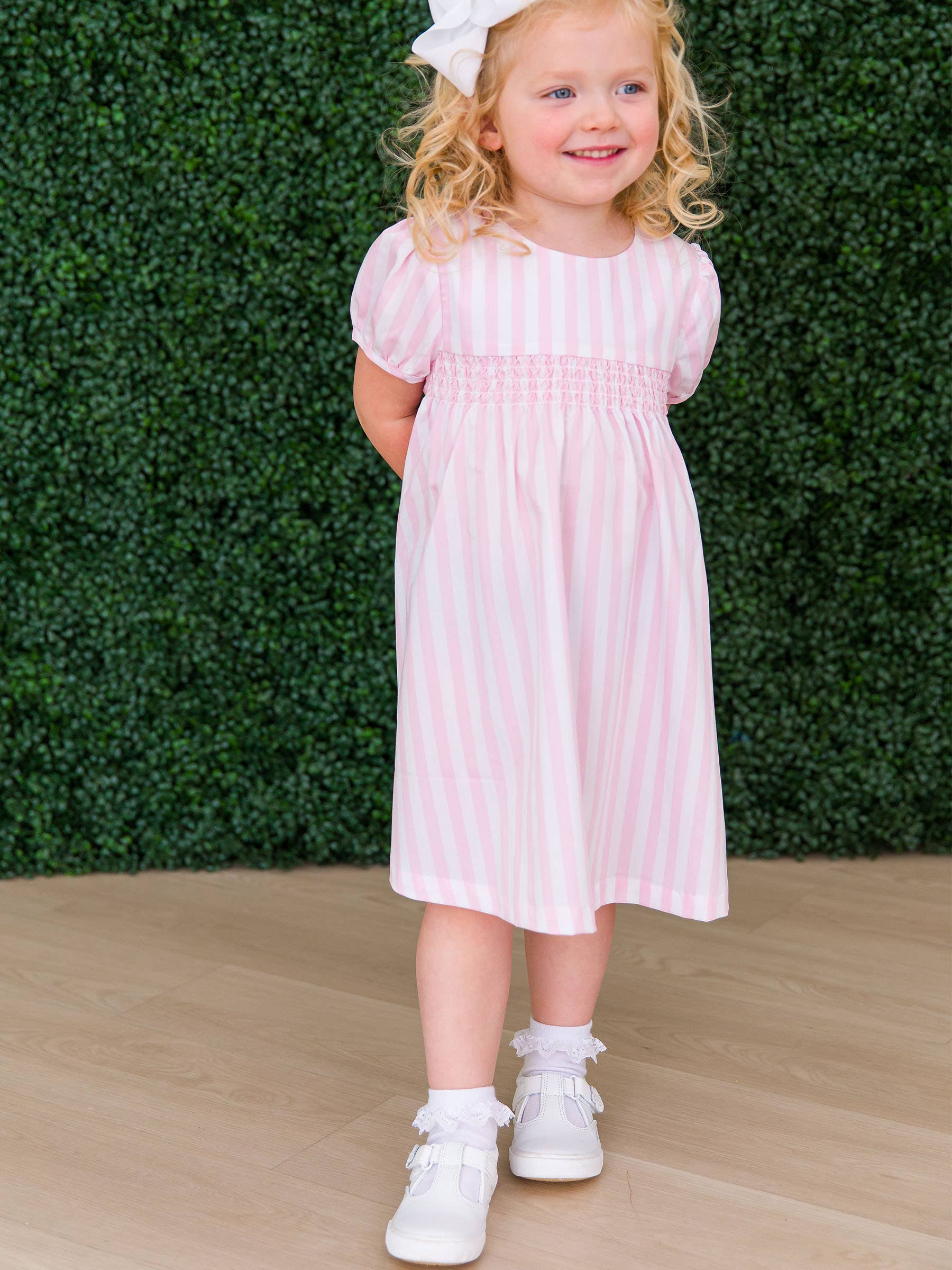 Young girl in a pink striped dress wearing white ruffled socks, standing against a green hedge.