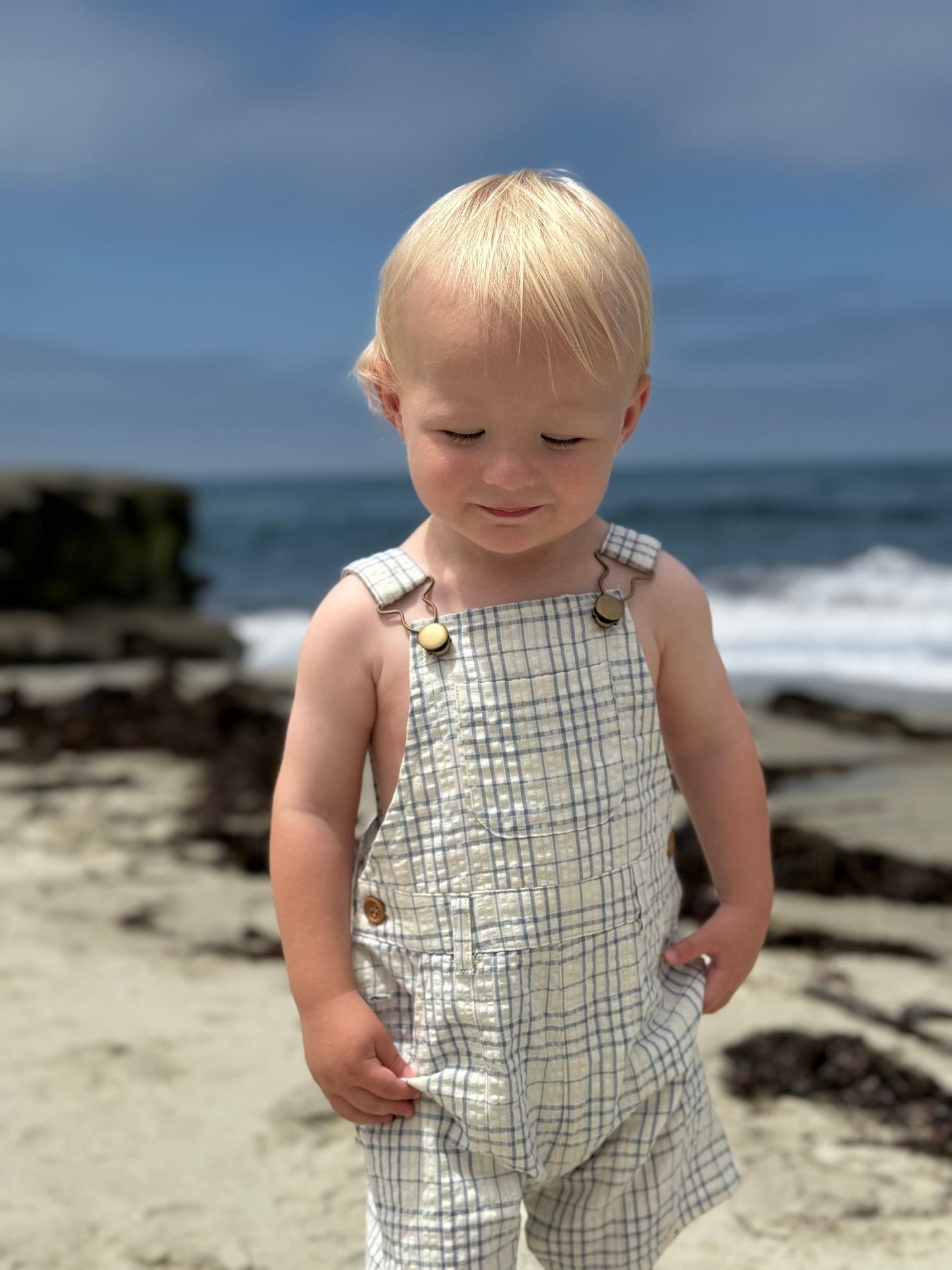 Child in plaid overalls standing on a beach with ocean in the background