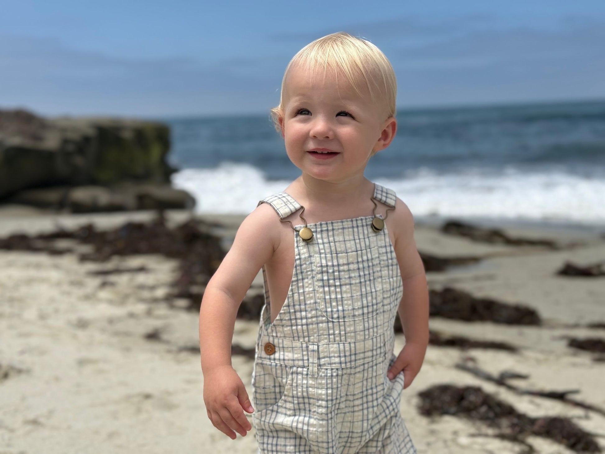 Child in plaid overalls standing on a beach with ocean waves in the background