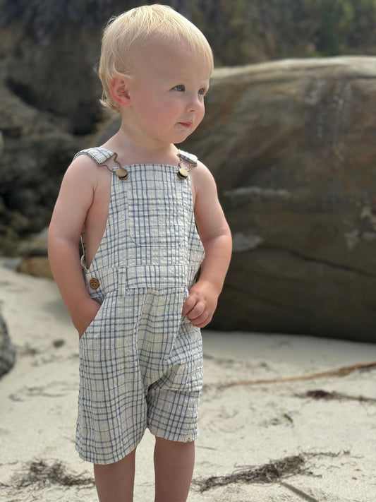 Child wearing a plaid romper standing on a rocky beach.