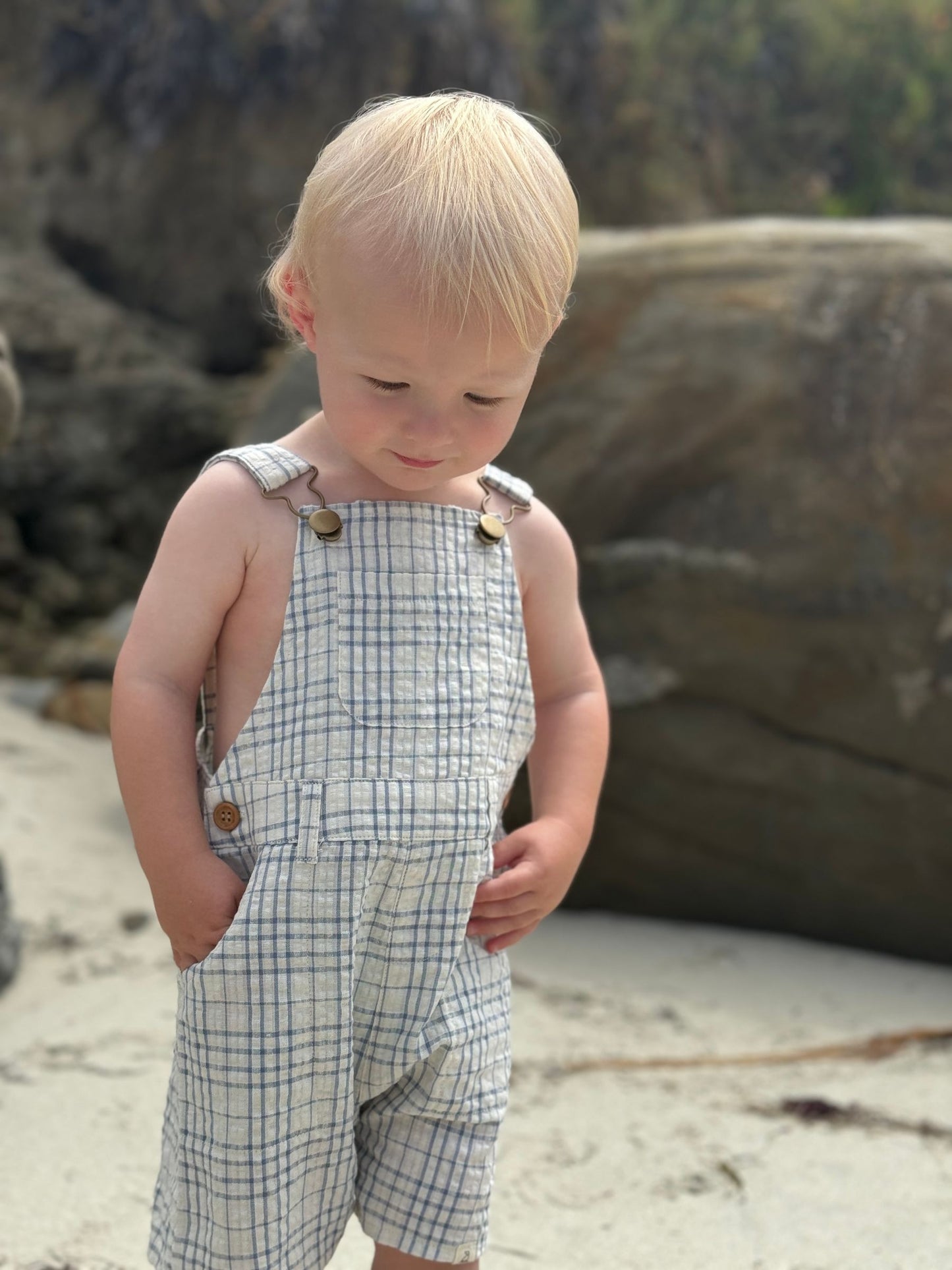 Child wearing a plaid romper standing on a rocky beach.