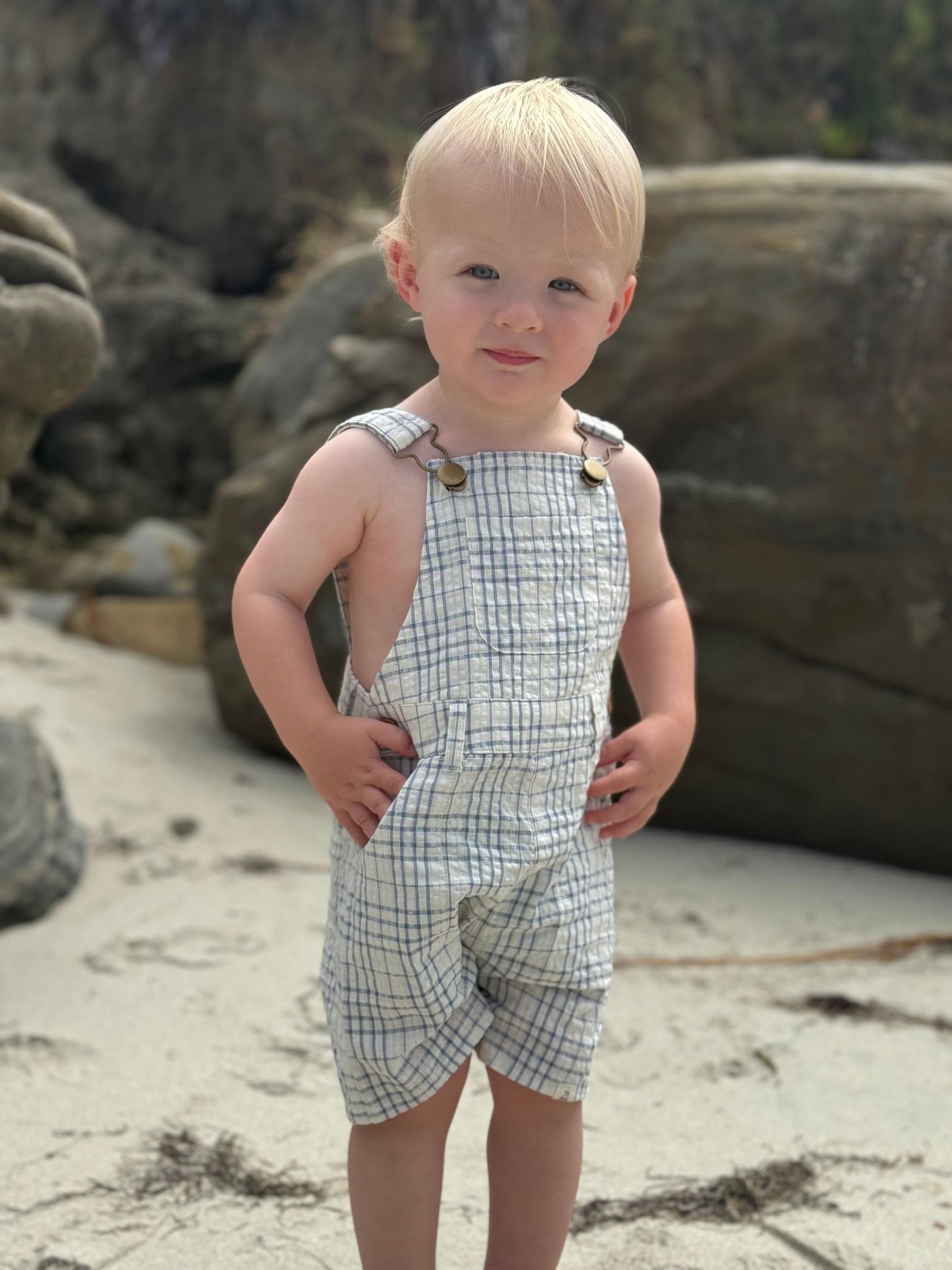 Child wearing plaid overalls standing on a rocky beach.