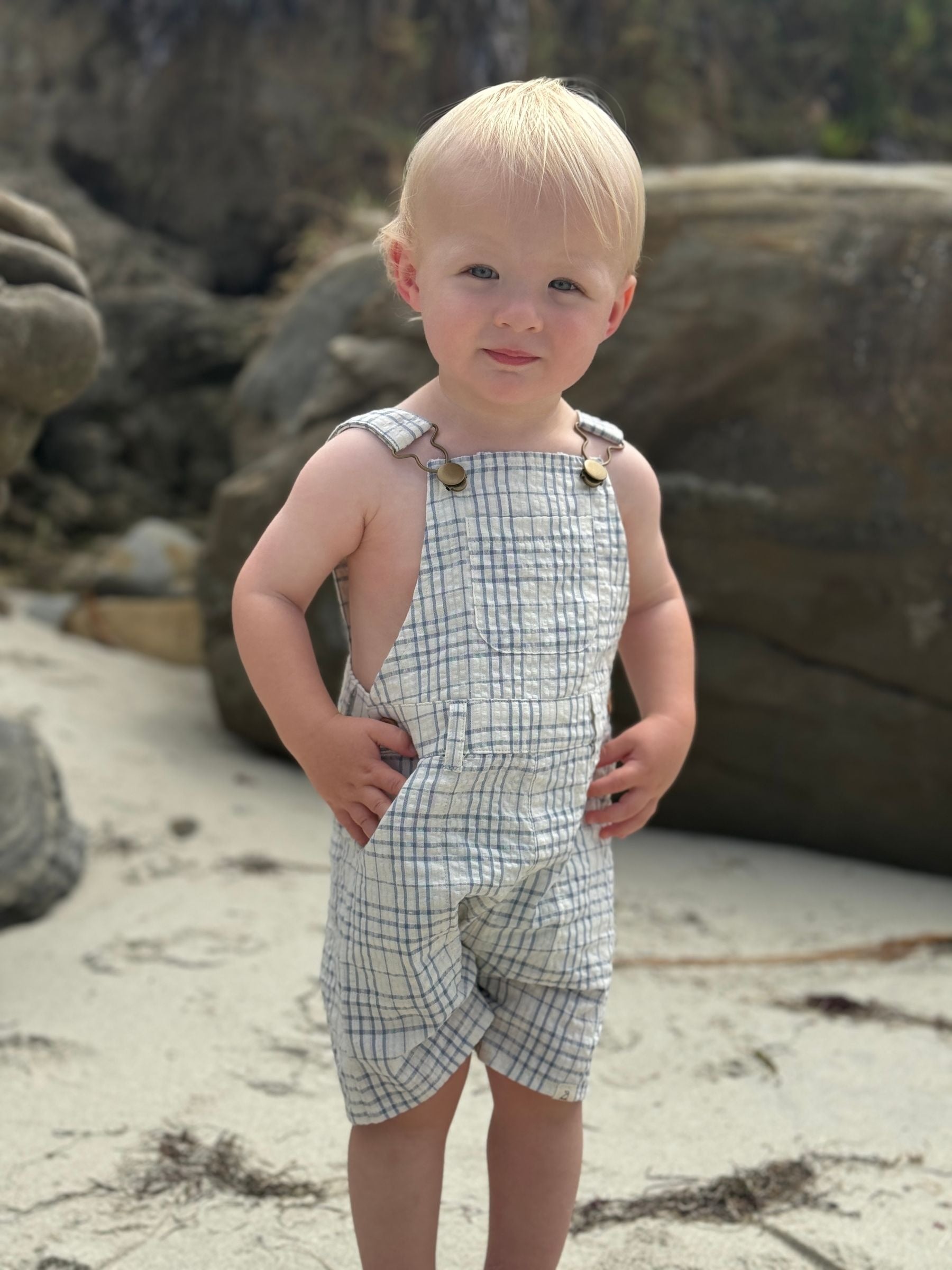 Child wearing plaid overalls standing on a rocky beach.