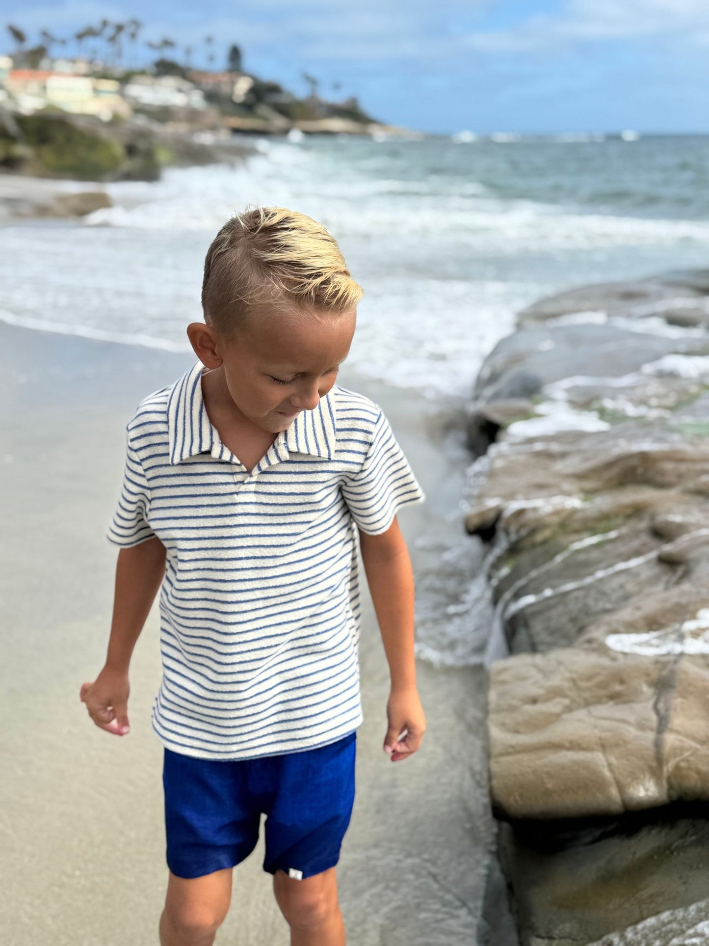 Child in a striped shirt and blue shorts standing on a beach with ocean waves in the background.