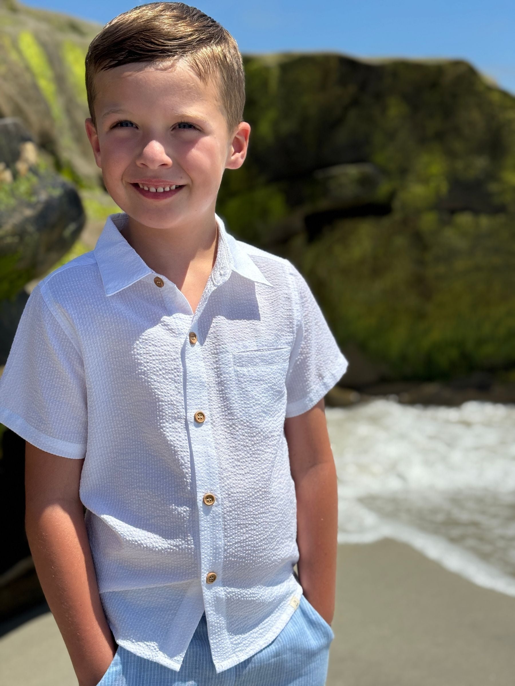 Young boy in a white shirt standing on a beach with greenery in the background