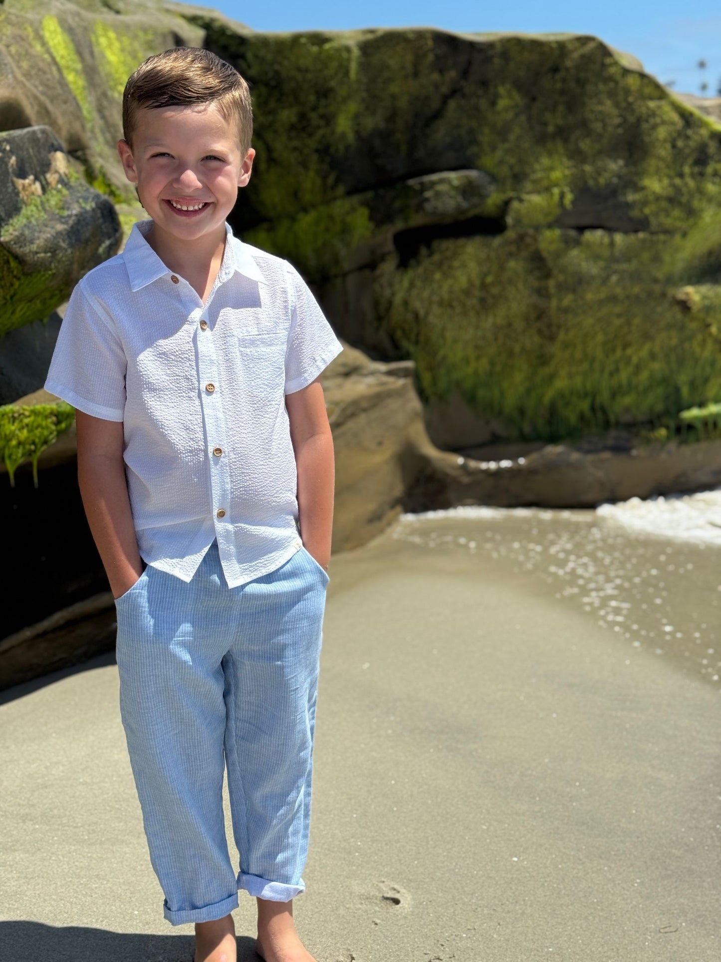 Young boy in a white shirt and light blue pants standing on a beach with large rocks in the background.