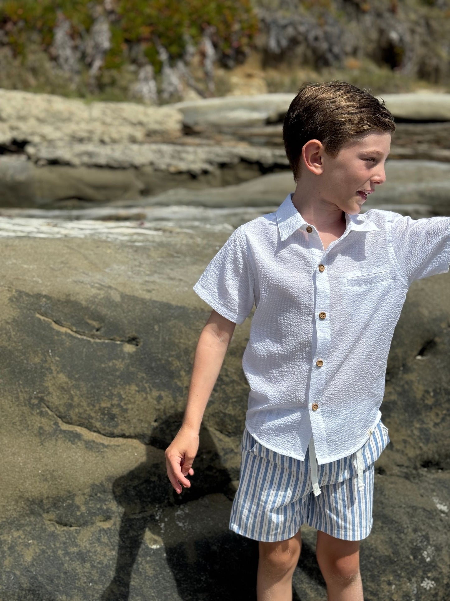 Child wearing a white shirt and striped shorts standing on a rocky beach.