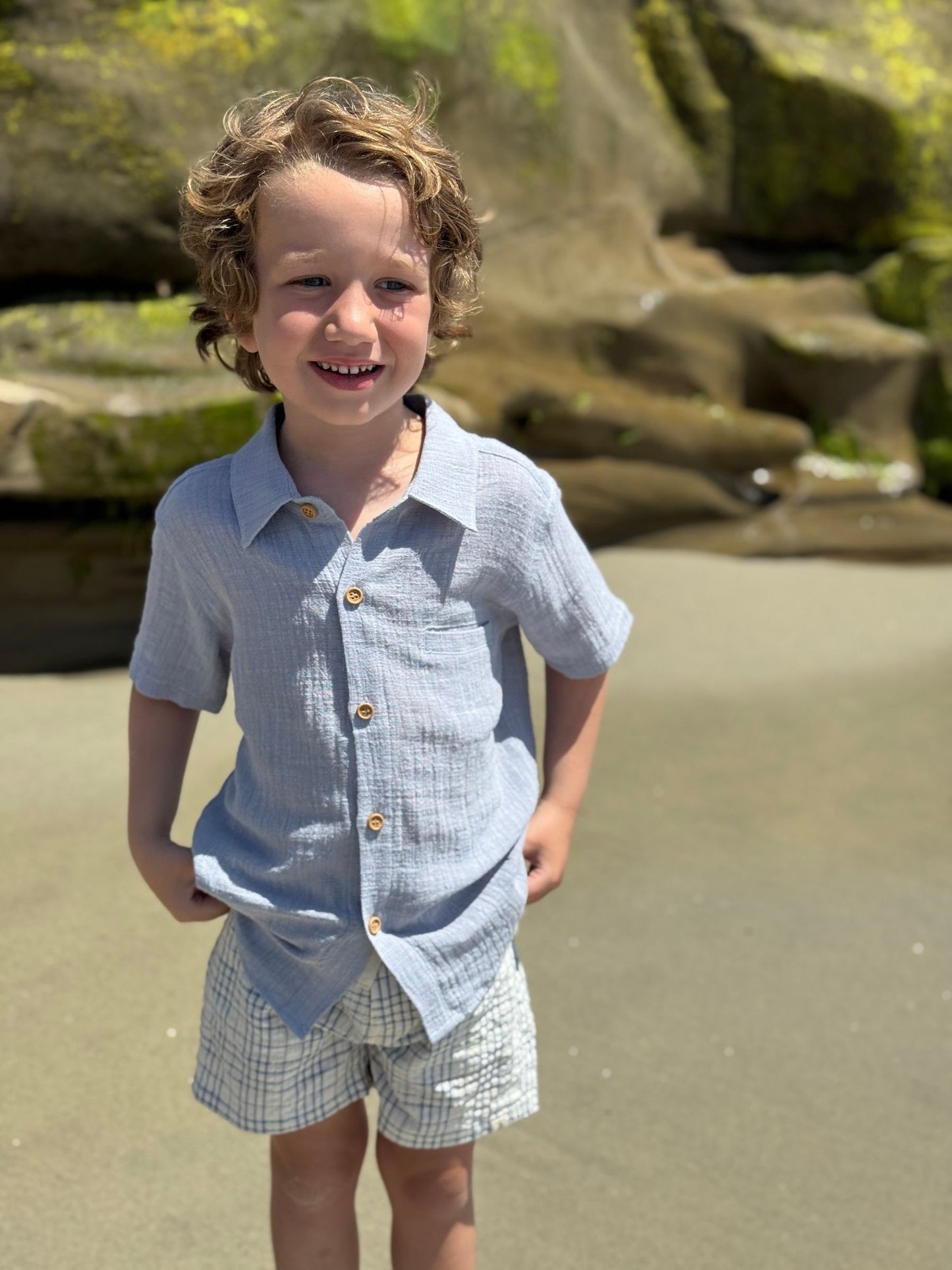Child wearing a light blue shirt with button details and white shorts on a sandy background.
