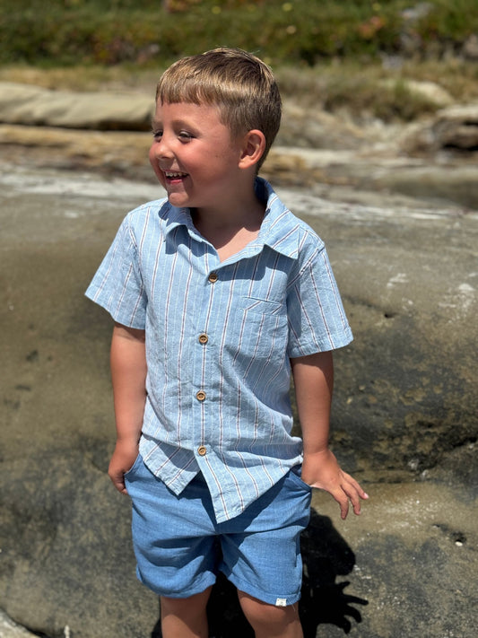 Young boy in a blue shirt and shorts standing on a rocky beach.