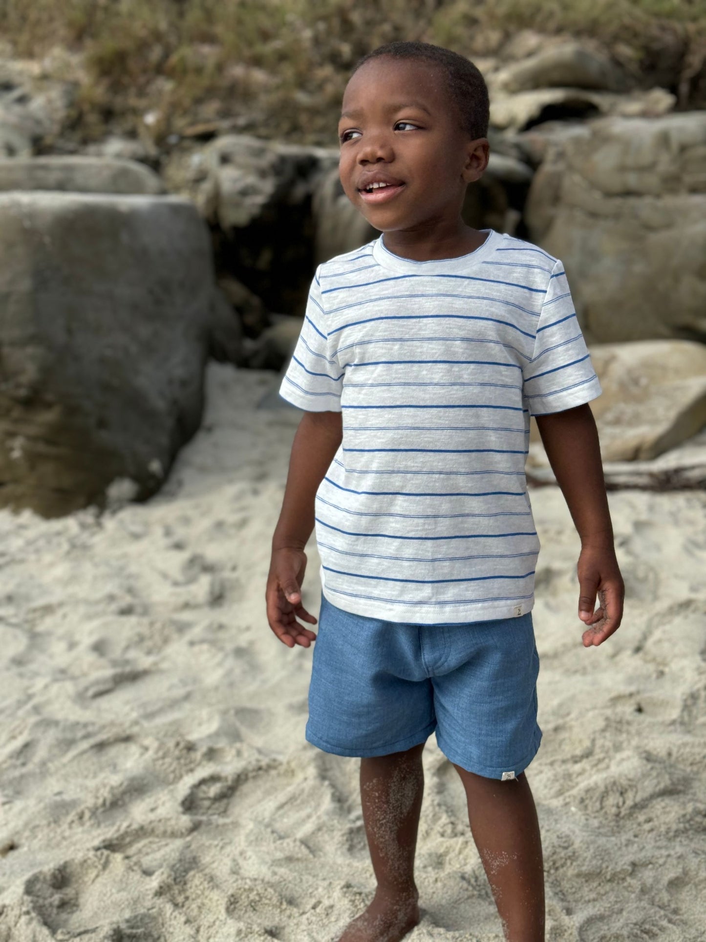 Child wearing a striped shirt and blue shorts standing on a sandy beach with rocks in the background