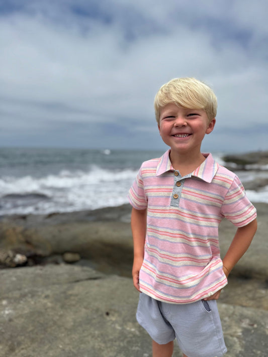 Young boy in a striped shirt standing on a rocky beach with ocean waves in the background.