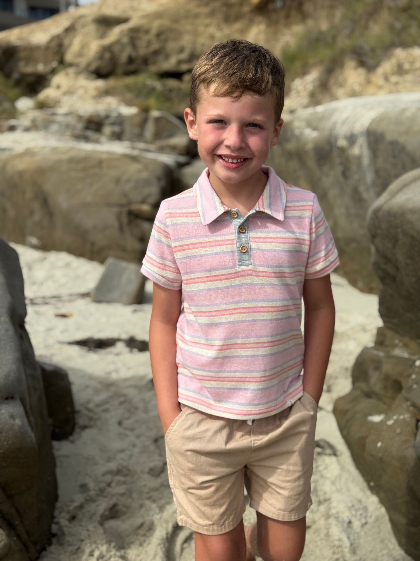 Young boy in a striped polo shirt and shorts standing on a rocky beach.