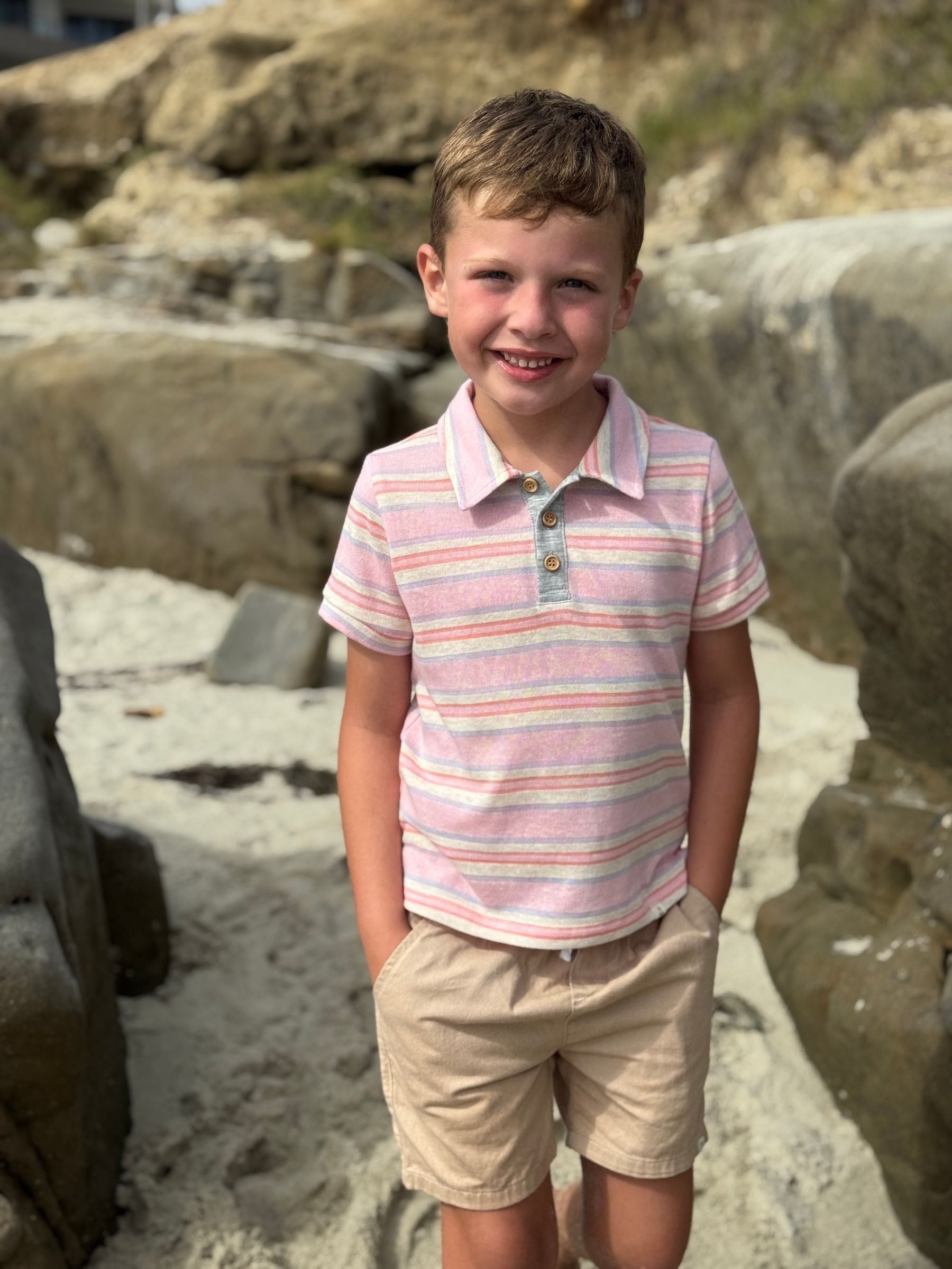 Young boy in a striped polo shirt and shorts standing on a rocky beach.