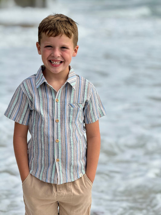 Young boy wearing a striped shirt and khaki pants standing by a body of water.