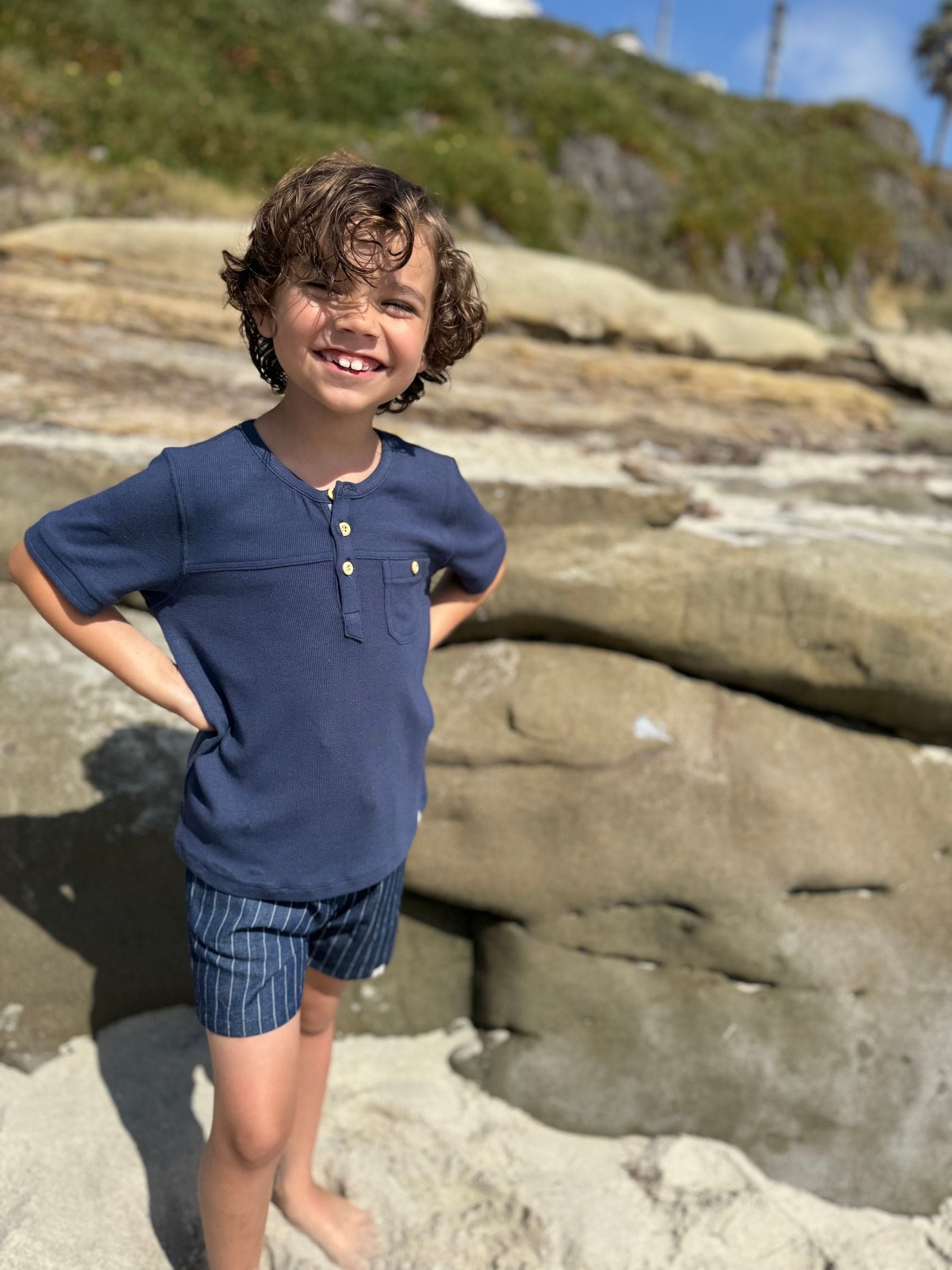 Child in a blue shirt and striped shorts standing on a rocky beach.