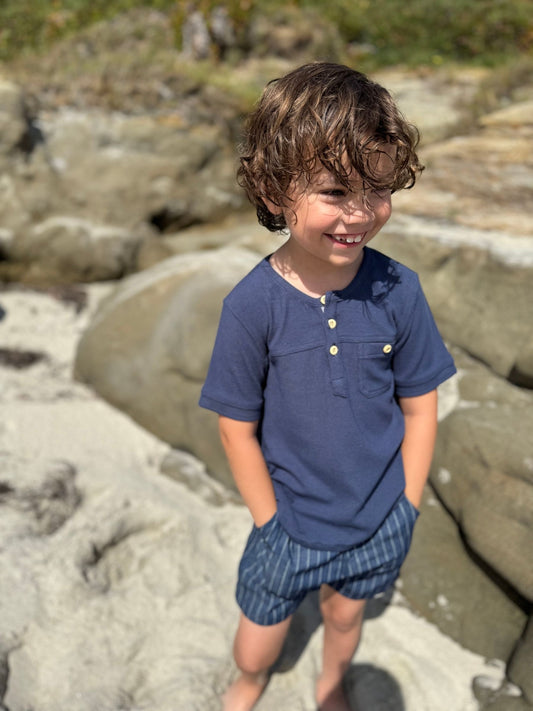 Child wearing a blue shirt and striped shorts standing on a rocky beach.