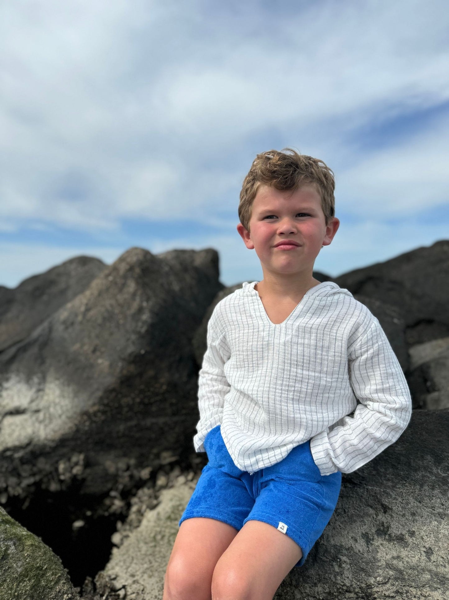 Young boy in a white sweater and blue shorts sitting on rocks with a cloudy sky background