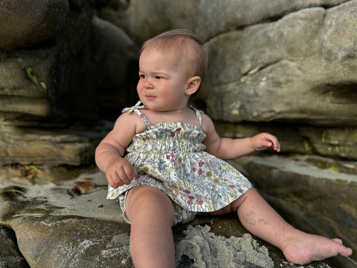 Baby in a floral dress sitting on rocks by a body of water