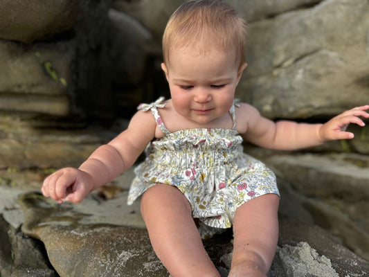 Baby in a floral outfit sitting on rocks by a body of water