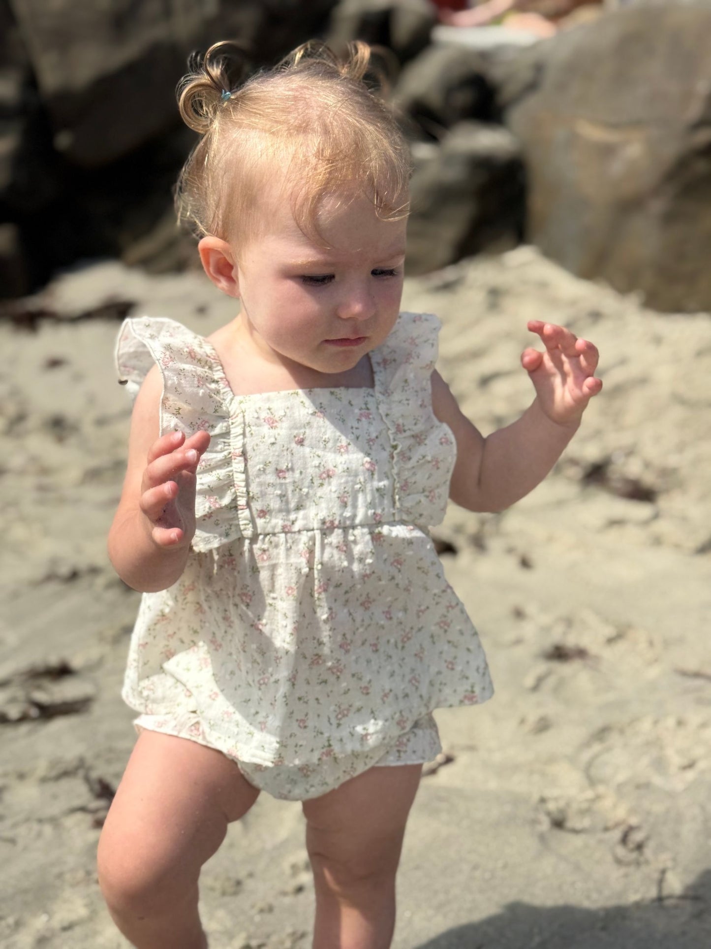 Young child in a floral dress standing on a sandy beach with rocks in the background
