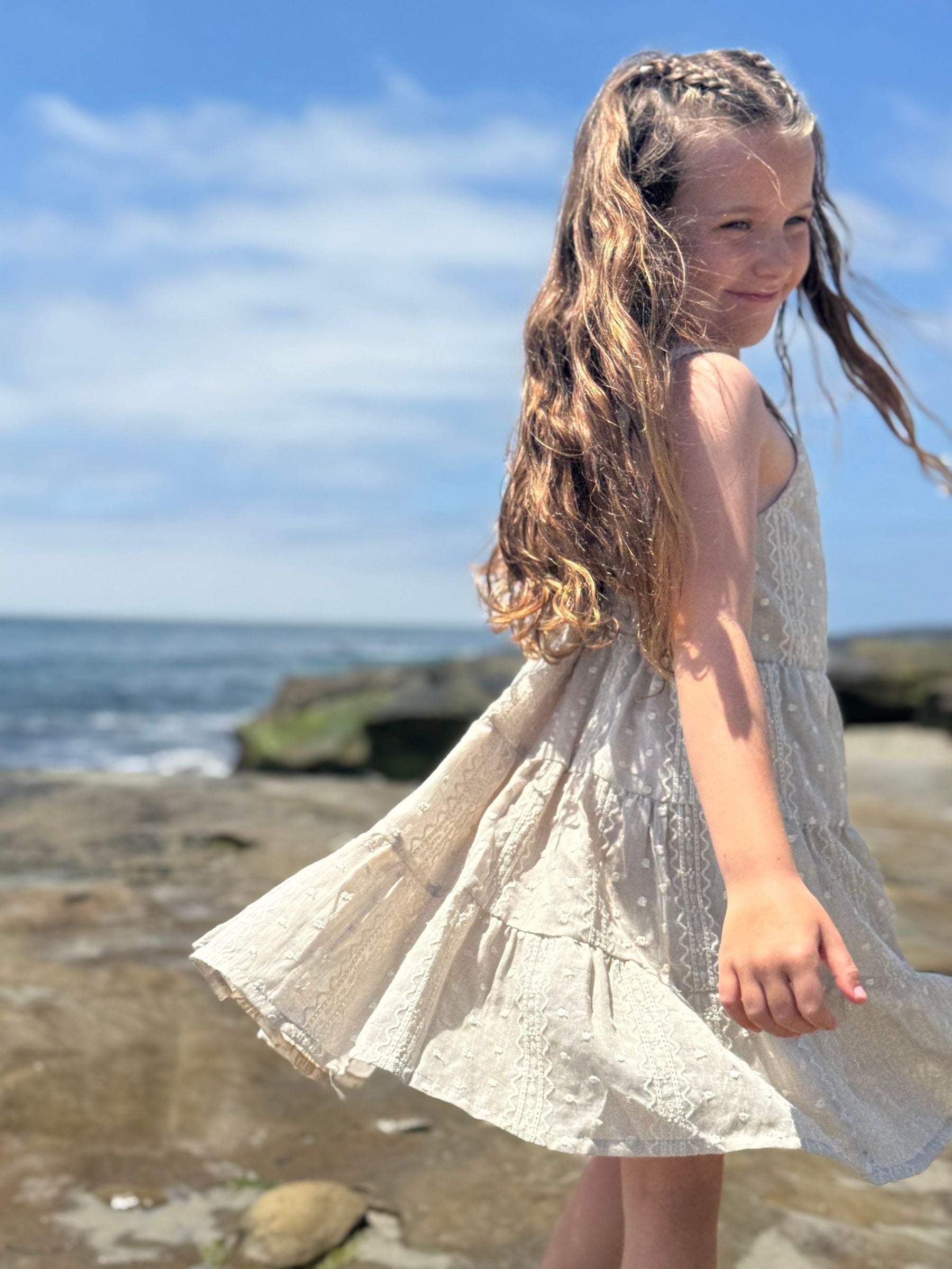 Young girl in a taupe dress standing on a rocky beach with ocean and sky in the background