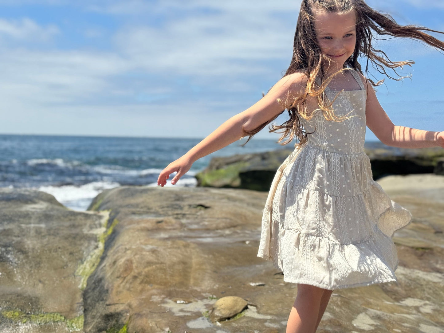 Young girl in a taupe dress standing on rocks by the ocean