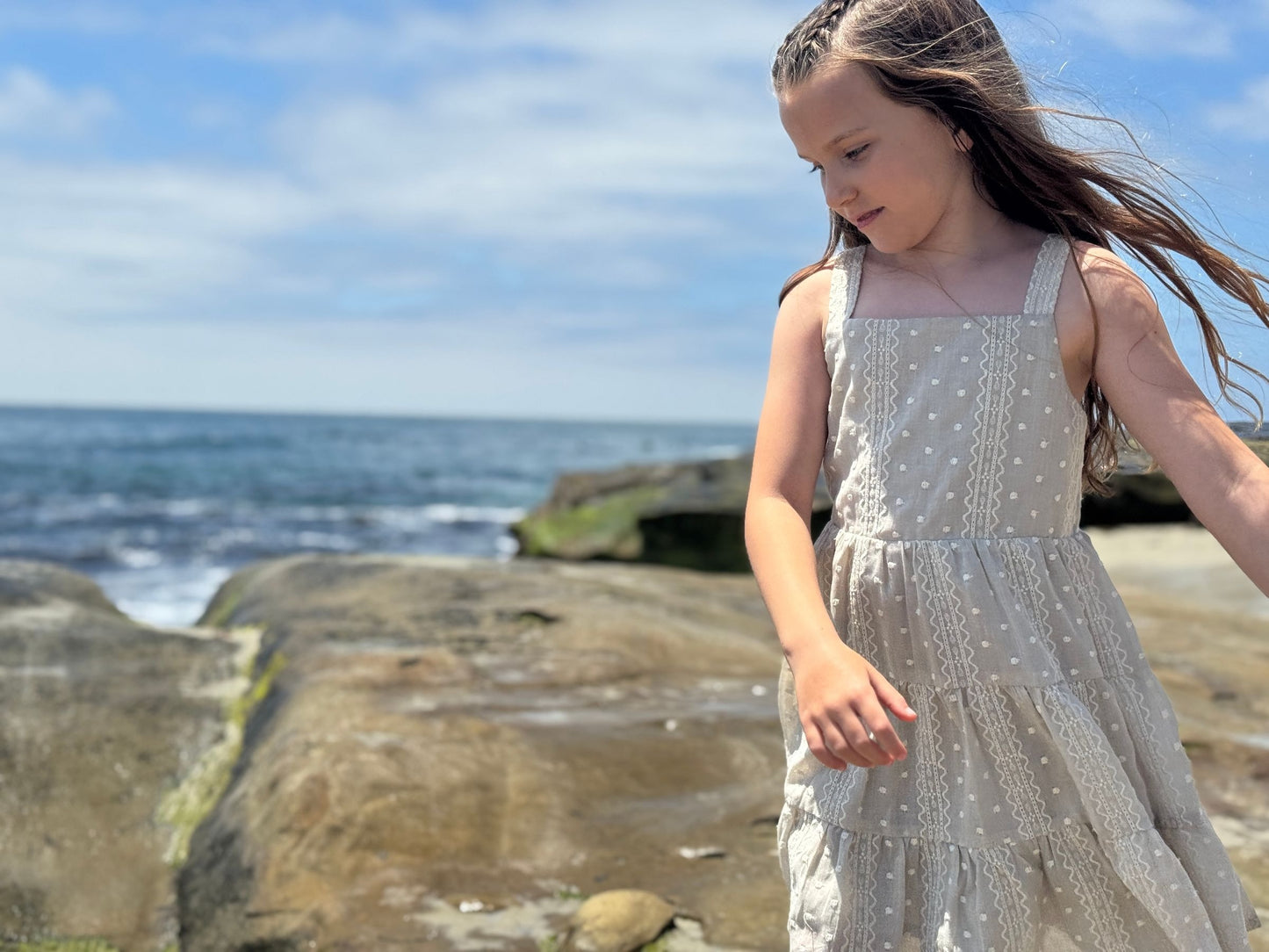 Young girl in a taupe dress standing on a rocky beach with ocean and sky in the background