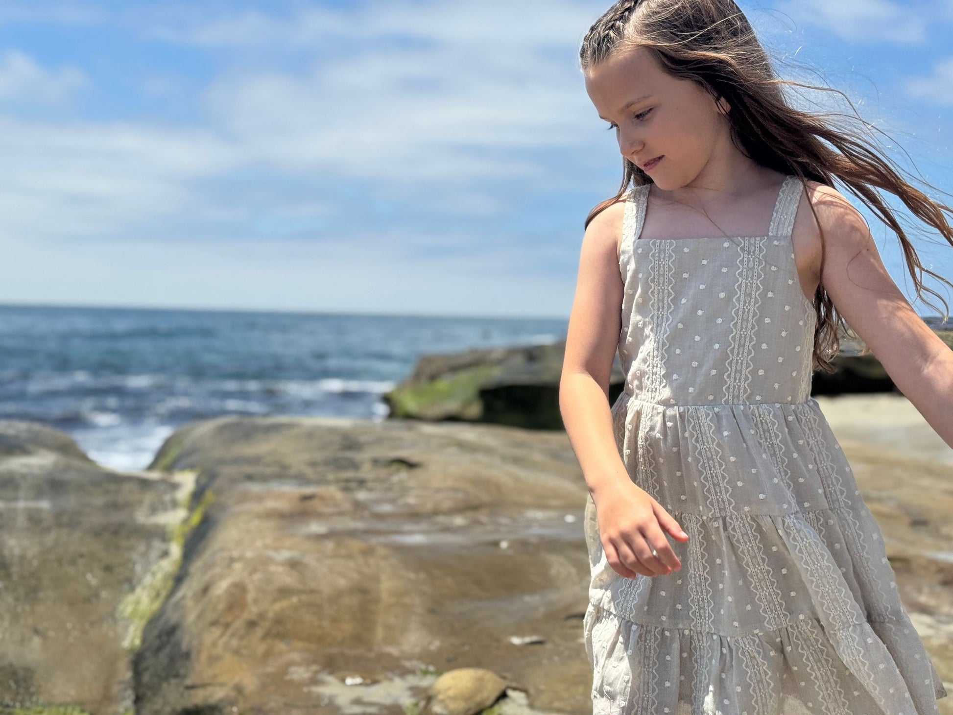 Young girl in a taupe dress standing on a rocky beach with ocean and sky in the background