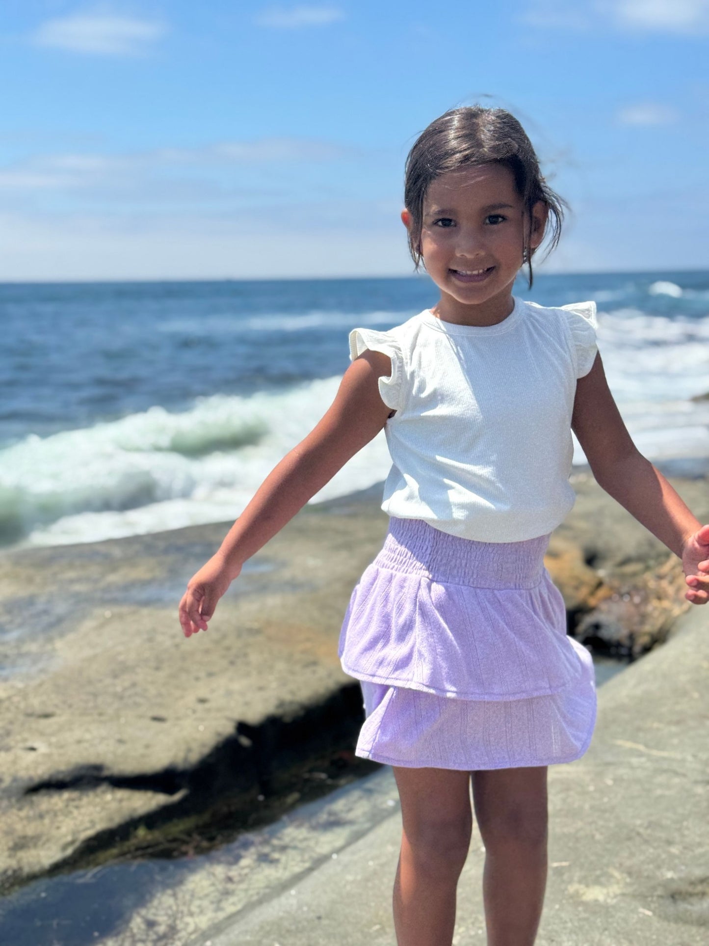 Young girl in a white top and purple skirt standing on a rocky beach with ocean waves in the background.