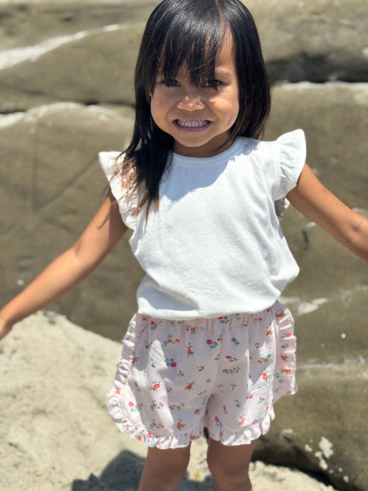 Young girl in a white top and floral shorts standing on a sandy beach.
