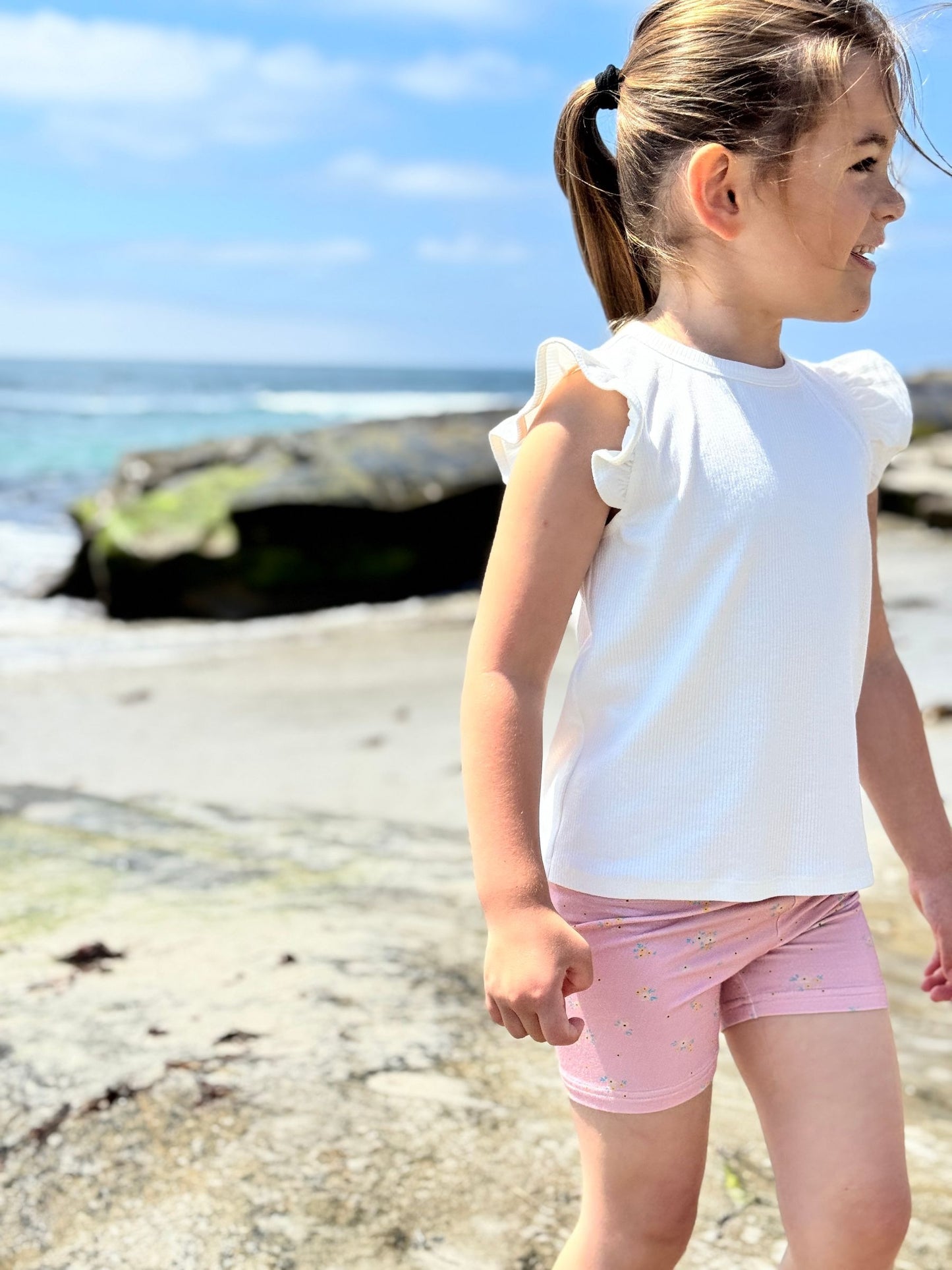 Young girl in a white top and pink shorts standing on a rocky beach.