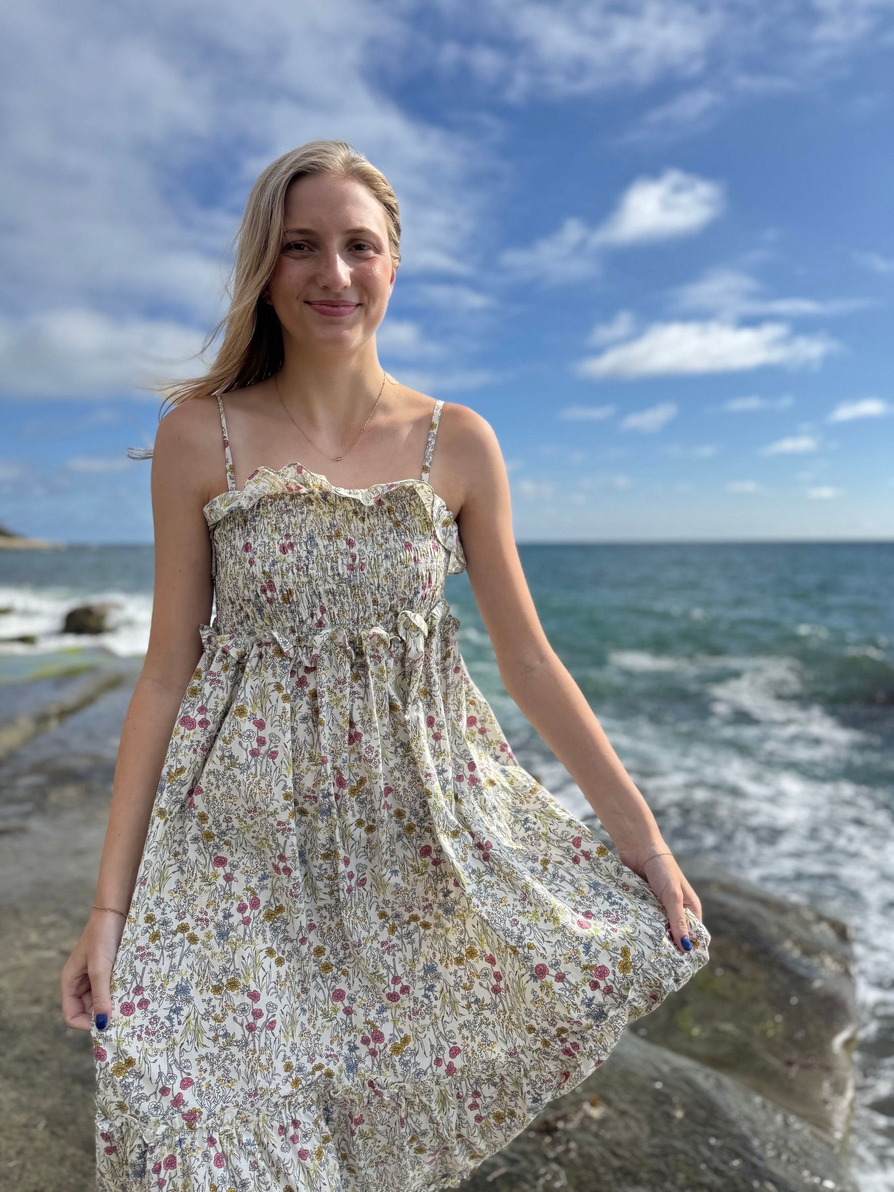 Woman in a floral dress standing on a rocky beach with ocean and sky in the background