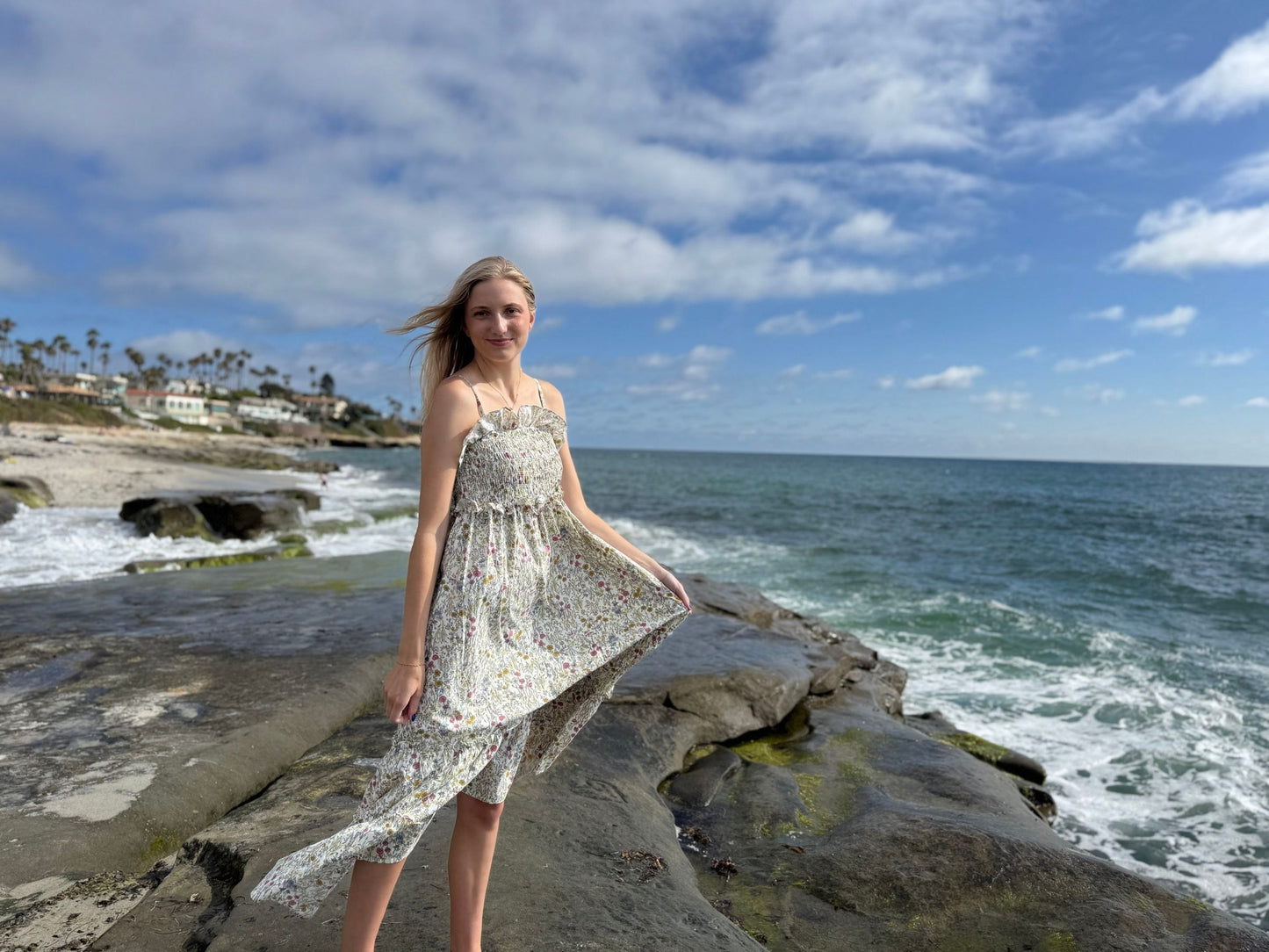 Woman in a dress standing on rocks by the ocean with a blue sky and clouds.