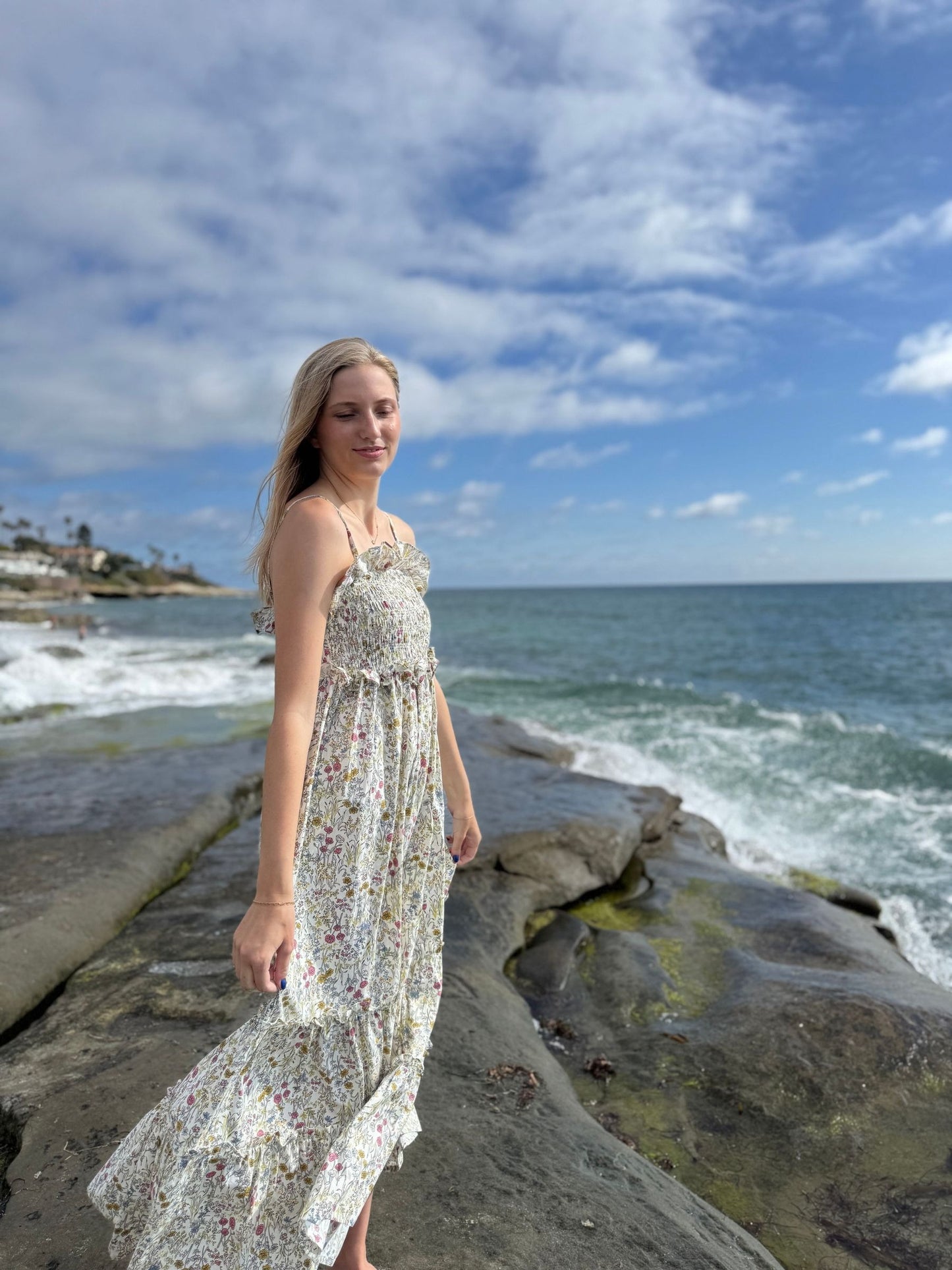 Woman in a floral dress standing on a rocky beach with ocean and sky in the background