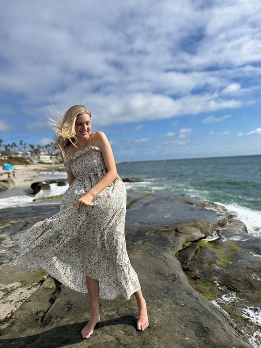 Woman in a floral dress standing on rocks by the ocean with a blue sky.
