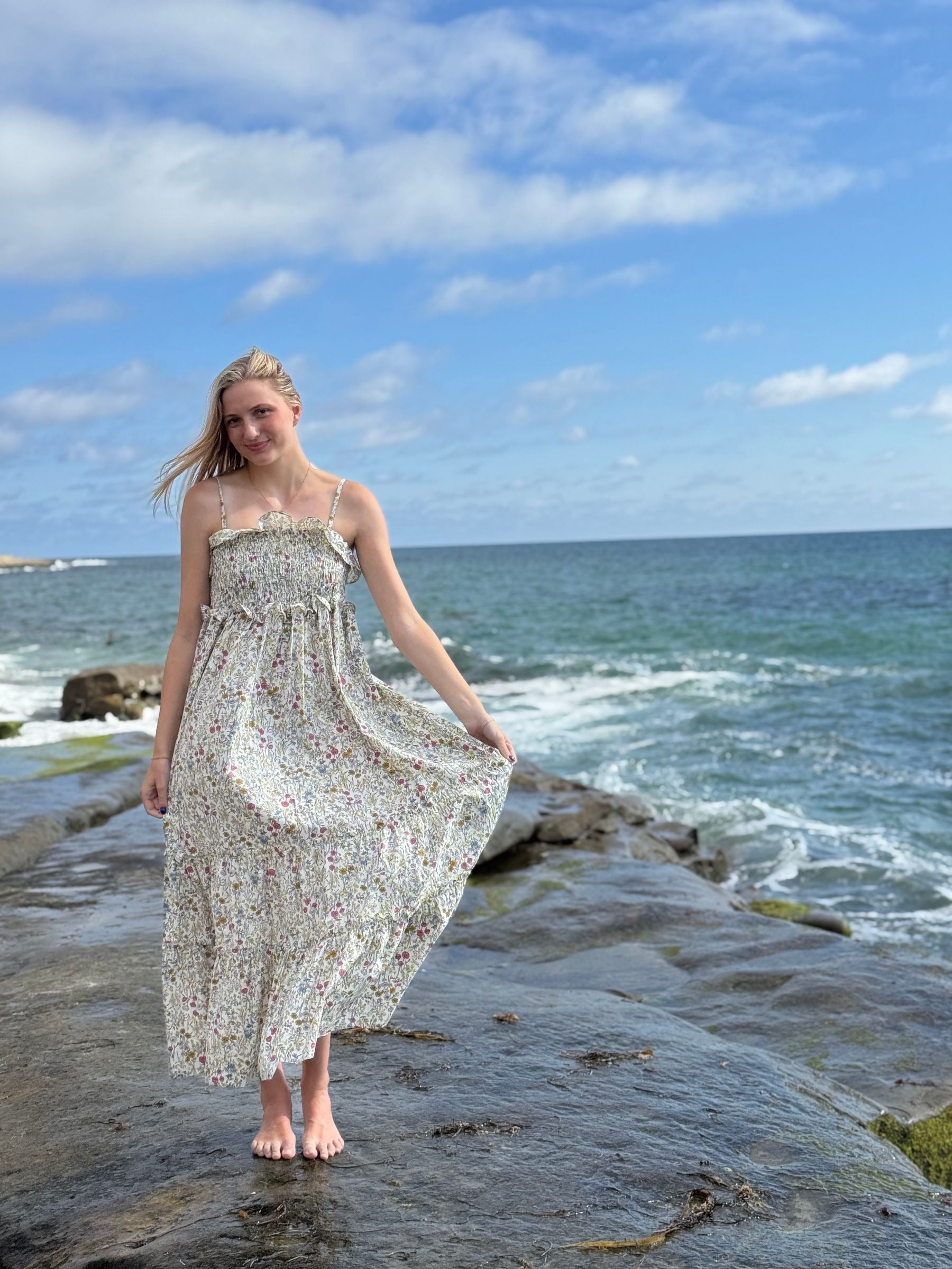 Woman in a floral dress standing on a rocky beach with ocean waves in the background