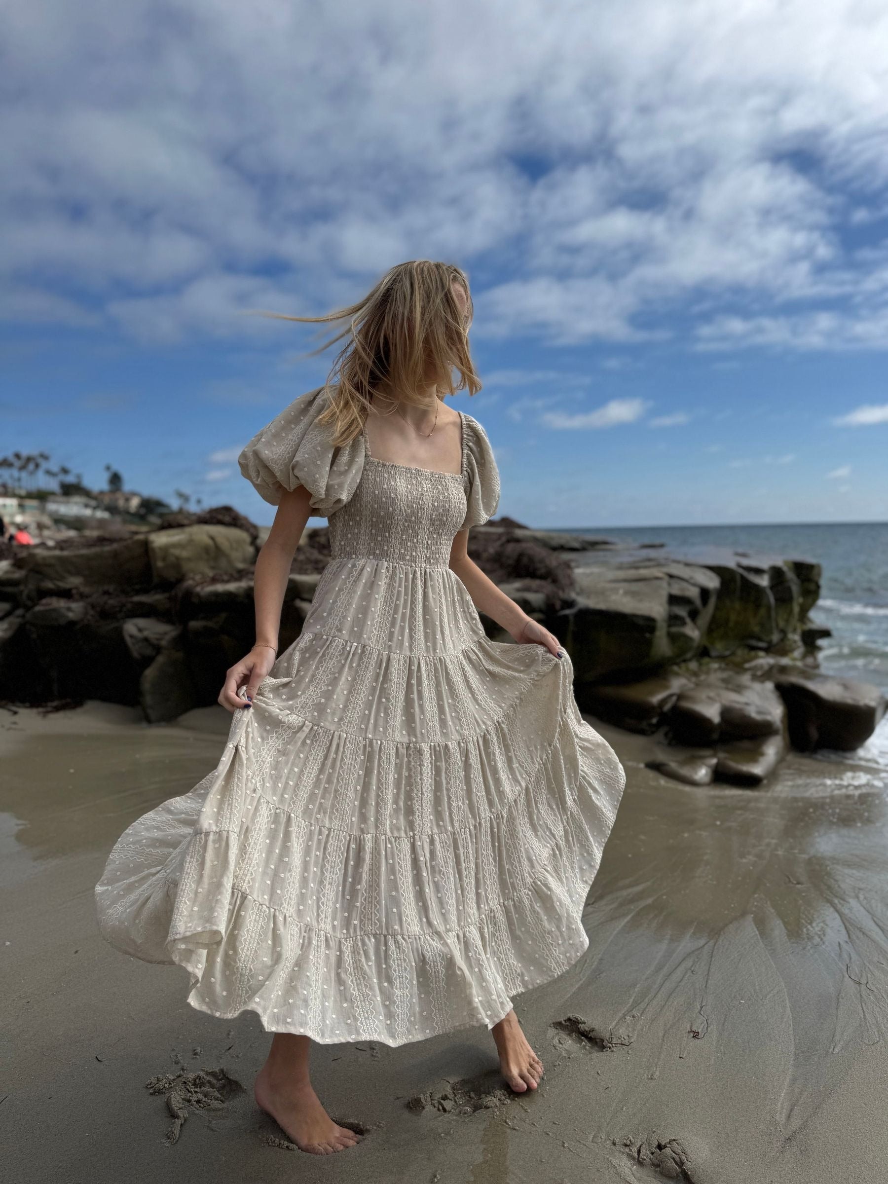 Woman in a flowing dress standing on a beach with rocky shoreline and blue sky.