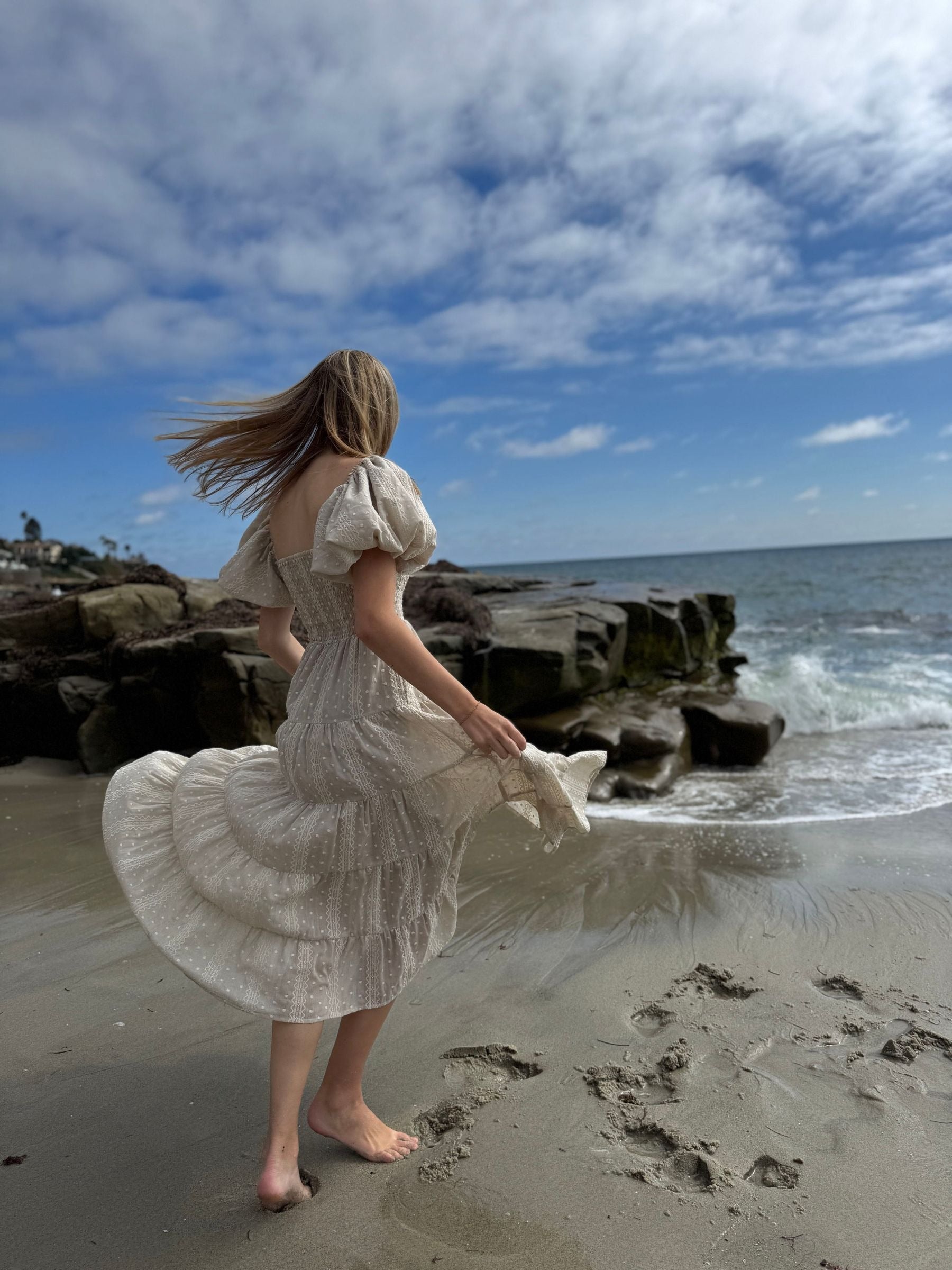 Woman in a white dress standing on a beach with rocky outcrops and ocean waves.