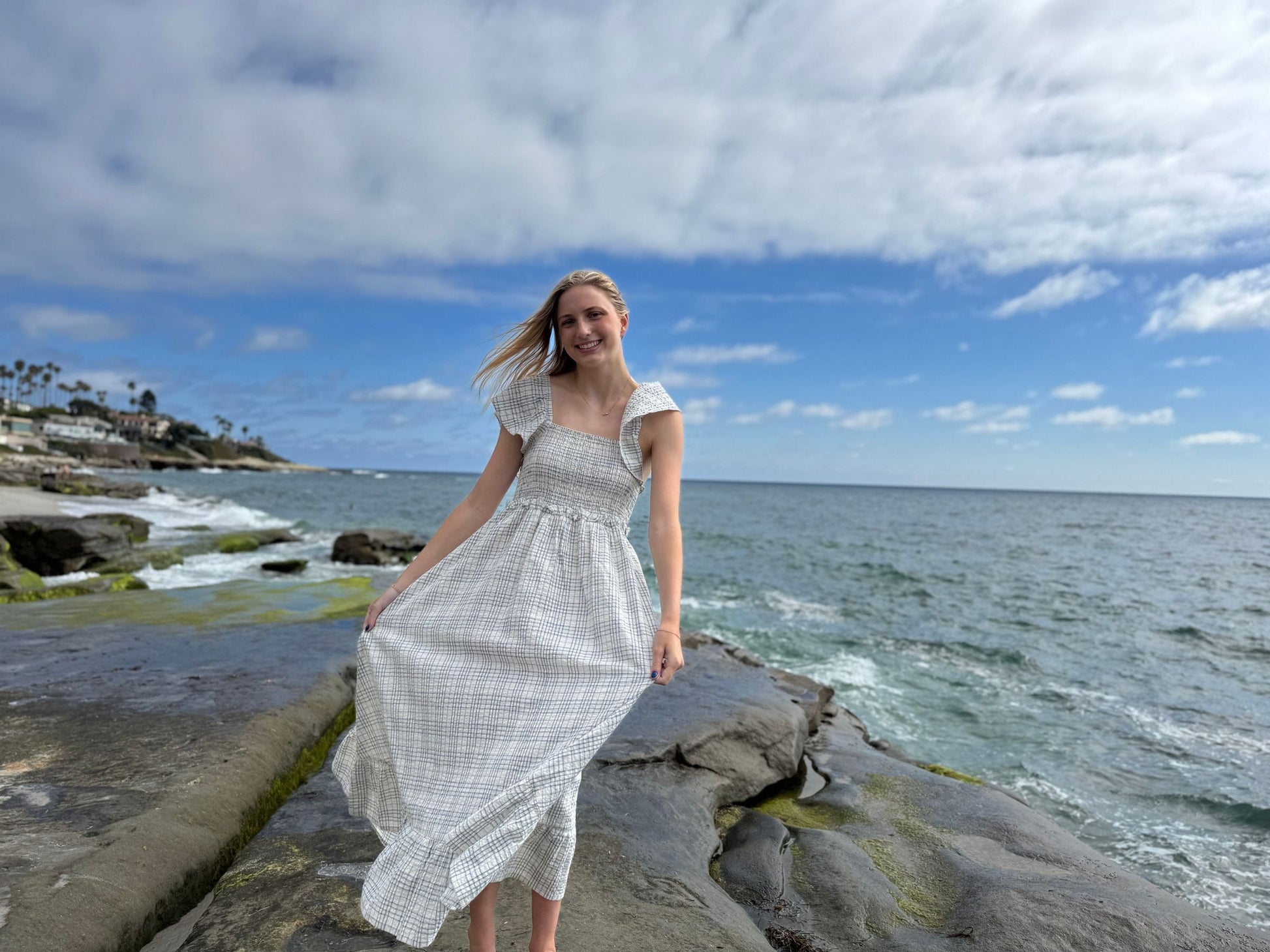 Woman in a white dress standing on rocks by the ocean with a blue sky and clouds.