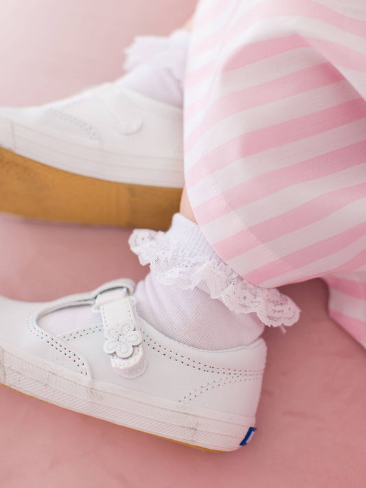 White ruffled socks worn with white sneakers with decorative laces worn by a child on a pink background